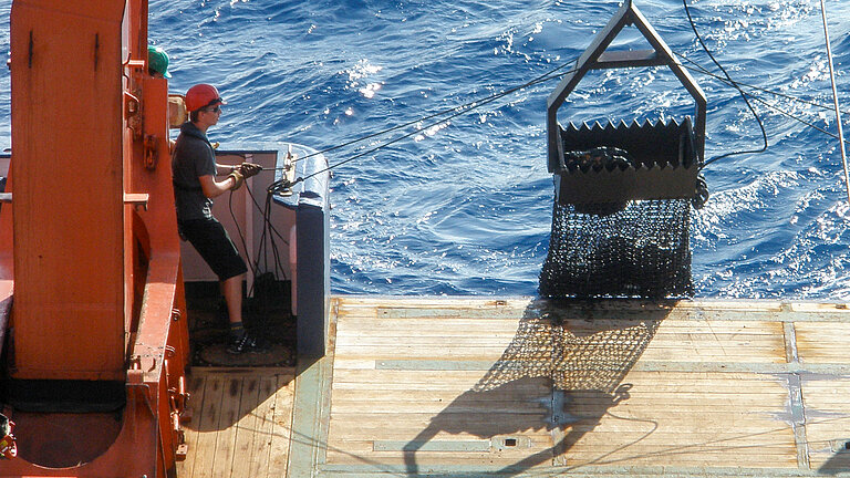 A young man wearing a red helmet stands at the stern of a ship and operates the crane. An iron sack filled with stones is pulled out of the water.