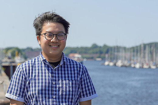 Jeng Hann Chong steht frontal im Bild und lacht. Im Hintergrund ist Wasser.