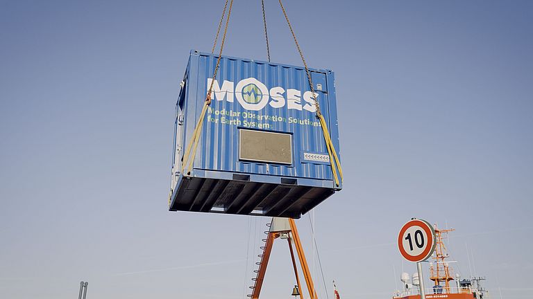 A dark blue container hangs in the air on the hook of a crane.