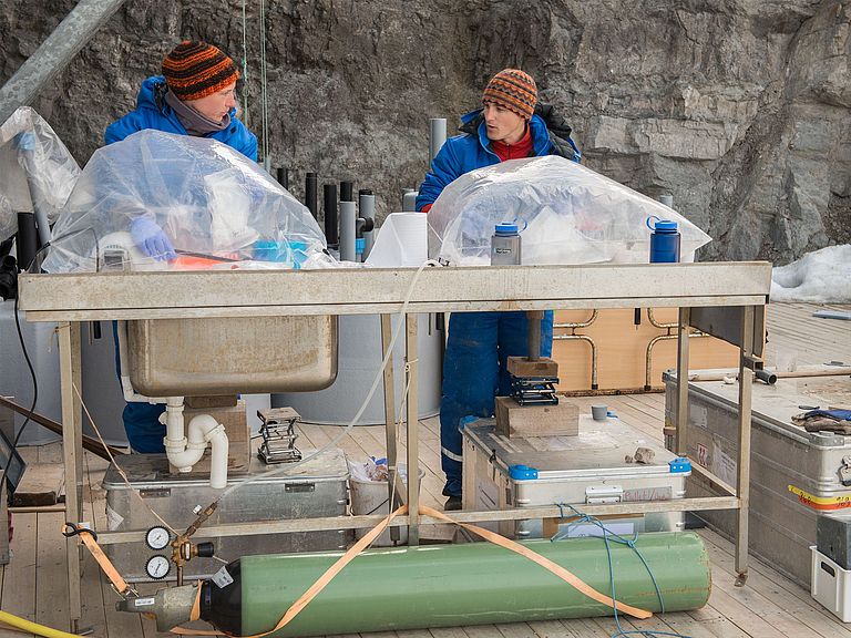A man and a woman in work clothes stand in front of a steep wall at a metal table and work on samples that are protected from the ambient air in large plastic bags.  Photo: Bo Barker Jørgensen