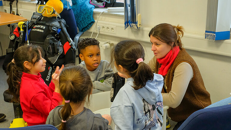 Eine junge Frau mit hellbraunen Haaren sitzt mit einigen Schulkindern im Kreis, im Hintergrund ist ein Taucheranzug zu sehen