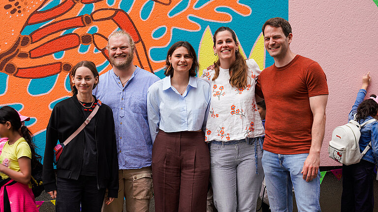 Three women and a man standing in front of a colourful painted wall and are smiling