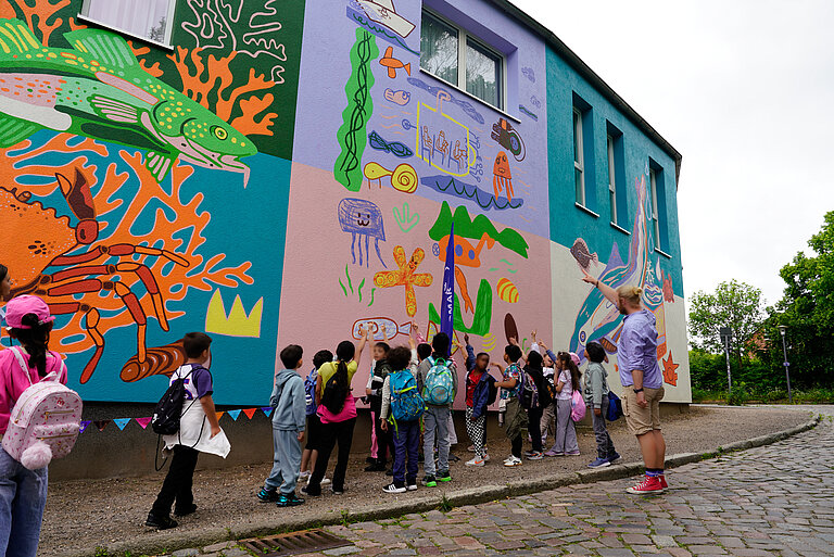 Primary school children are standing in front of a painted wall and pointing at the pictures