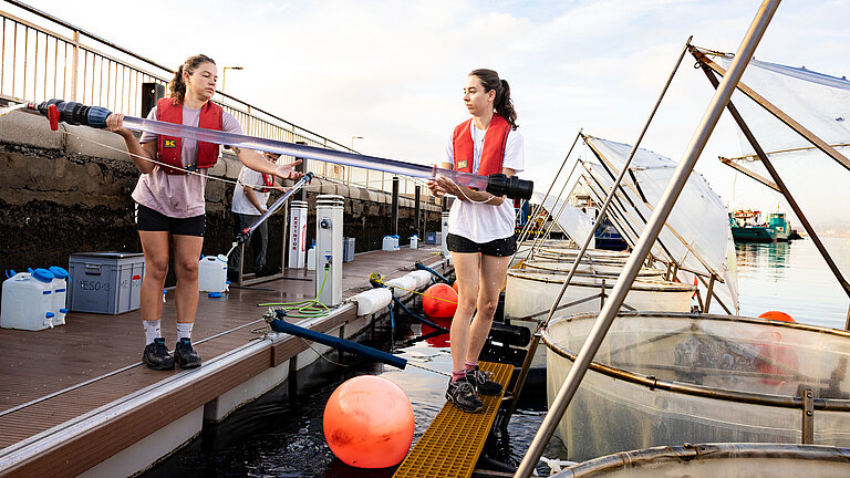 Am Kai im Hafen stehen zwei junge Frauen mit Schwimmwesten, im Wasser vor ihnen sind Kunststoffringe mit regenschirmförmigen Deckeln aus transparentem Material. Die beiden Frauen haben ein langes Rohr mit einer transparenten Flüssigkeit in den Händen. 