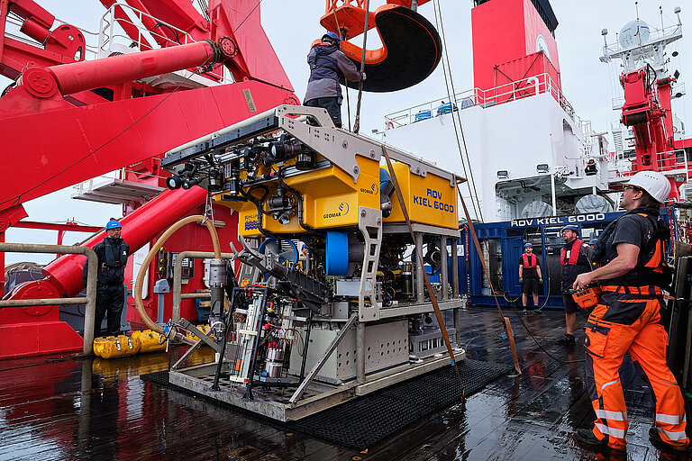 A large yellow piece of research equipment – a diving robot – is standing on the deck of a ship, with a few technicians wearing helmets standing next to it