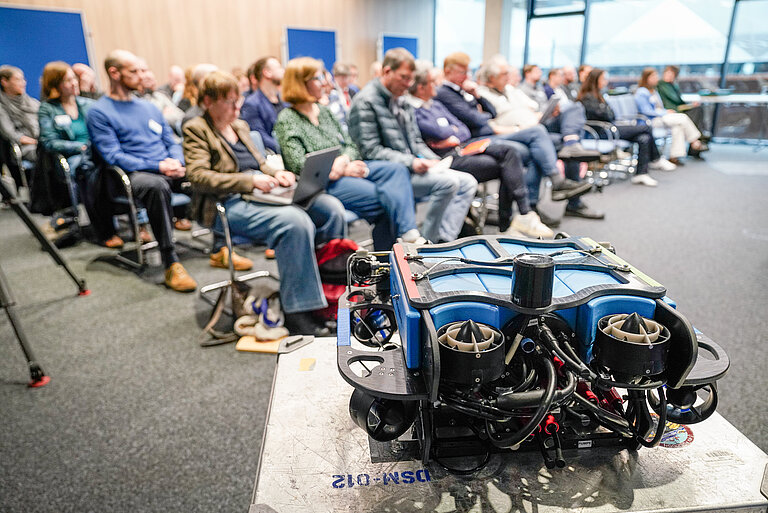 [Translate to English:] Im Vordergrund ein meereswissenschaftliches Grät, unscharf dahinter ein Konferenzraum, mit Publikum gefüllt
