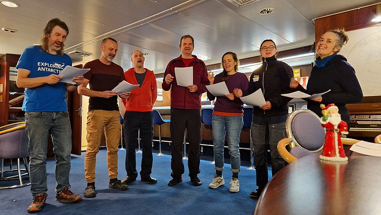 Seven men and women stand in a low room aboard a ship and sing.