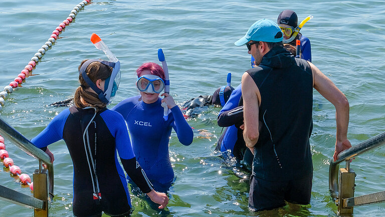 A young man wearing a cap and sunglasses and a few children are standing on a flight of steps leading down into the water; the children are wearing wetsuits.