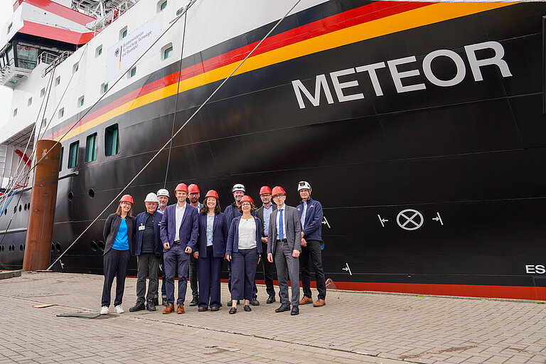 Eine Gruppe Menschen vor der hohen schwarzen Bordwand eines Schiffes im Hafen. Auf dem Bug ist der Schriftzug "METEOR" zu lesen.