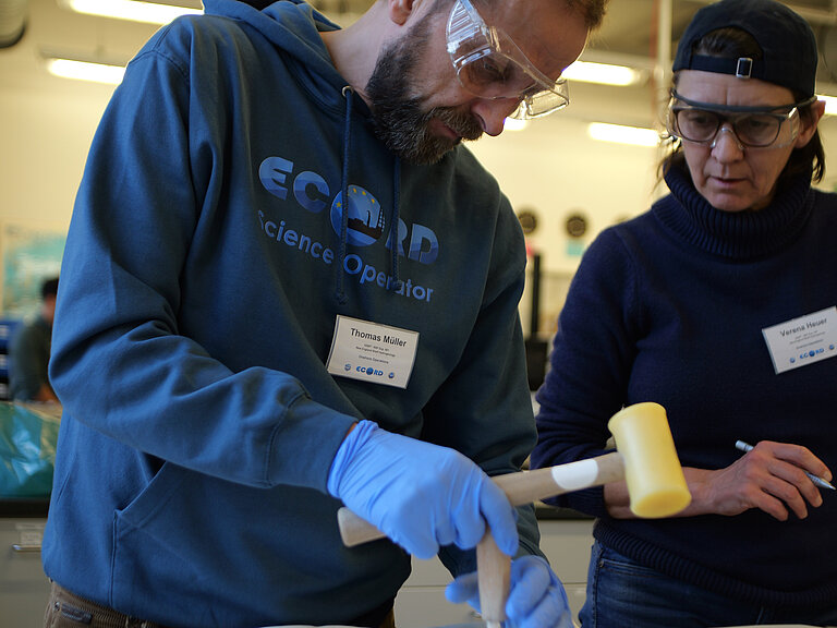 Two people, a man and a woman. The man is wearing transparent laboratory safety goggles and is working on a rock lying on the table with a chisel and a plastic hammer.