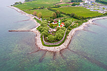 An aerial view of a peninsula jutting out into the sea, with star-shaped groynes. On the peninsula stands a green-and-white-striped lighthouse; green meadows and a sewage treatment works can be seen in the background.