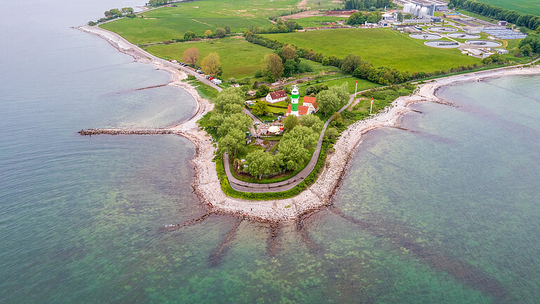 Luftaufnahme einer Halbinsel, die mit sternförmigen Buhnen ins Meer ragt. Auf der Insel steht ein grün-weiß gestreifter Leuchtturm, im Hintergrund sind grüne Wiesen zu sehen und ein Klärwerk.