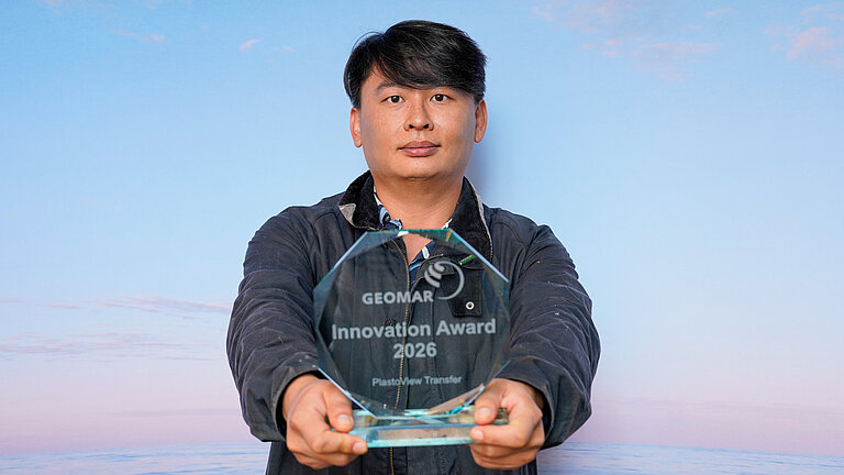 Ein junger Mann mit schwarzen Haaren und asiatischen Gesichtszüge hält einen gläsernen, gravierten Preis in die Kamera.