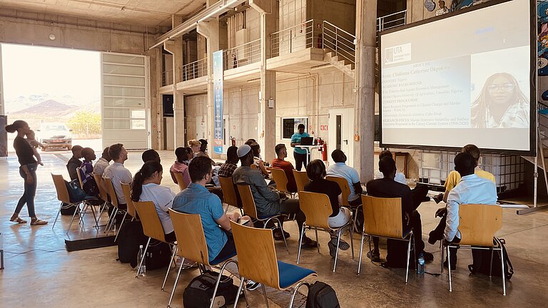 Young people of colour are sitting in front of a large screen in the sun-drenched hangar of a research institute