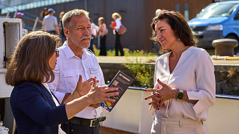 Two women in elegant trouser suits and a man in full dress uniform on a ship, with a tall building with a shiny silvery façade in the background.