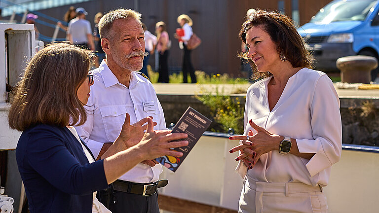 Zwei Frauen und ein Mann in Paradeuniform auf einem Schiff, im Hintergrund ein hohes Gebäude mit Silberfarben glänzender Fassade