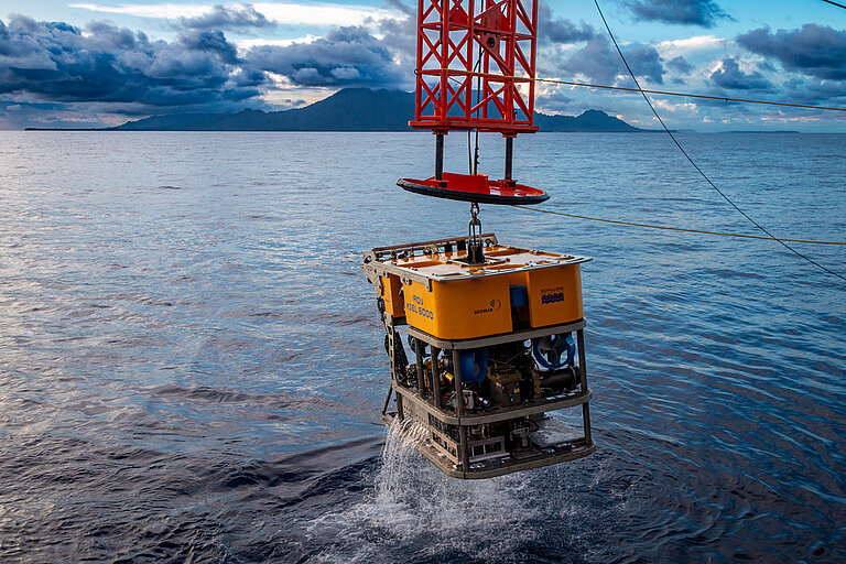 Ein gelber eckiger Tauchroboter wird an einem roten Kran ins Wasser gelassen.
