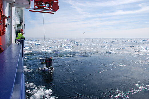 A CTD (conductivity, temperature, depth) sampling frameis lowered into the sea from aboard a research vessel. Ice floes can be seen on the water.