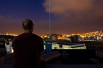A green laser goes from the OSCM vertically into the night sky over Mindelo. In the foreground a man looks in the direction of the laser.