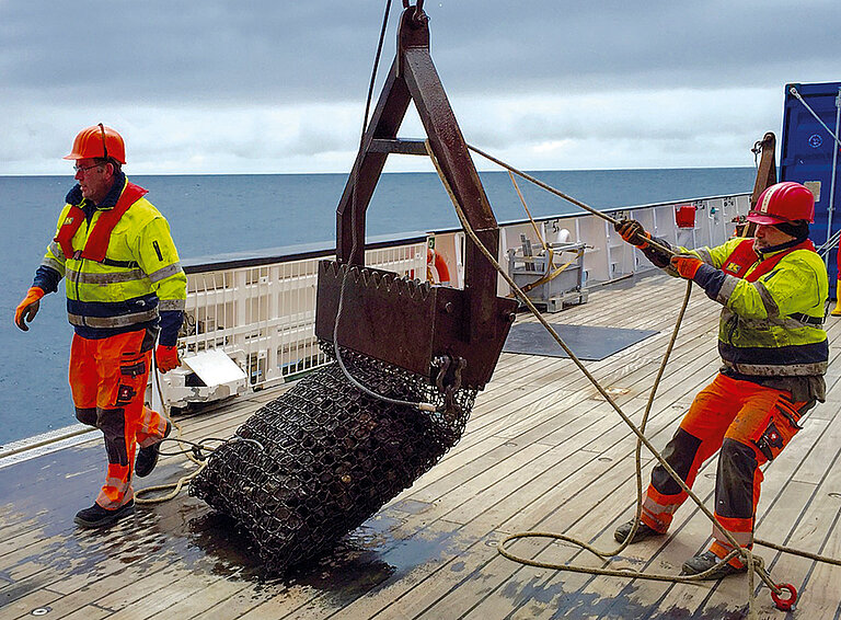 A dredge is returned to the research vessel by two men wearing safety helmets.