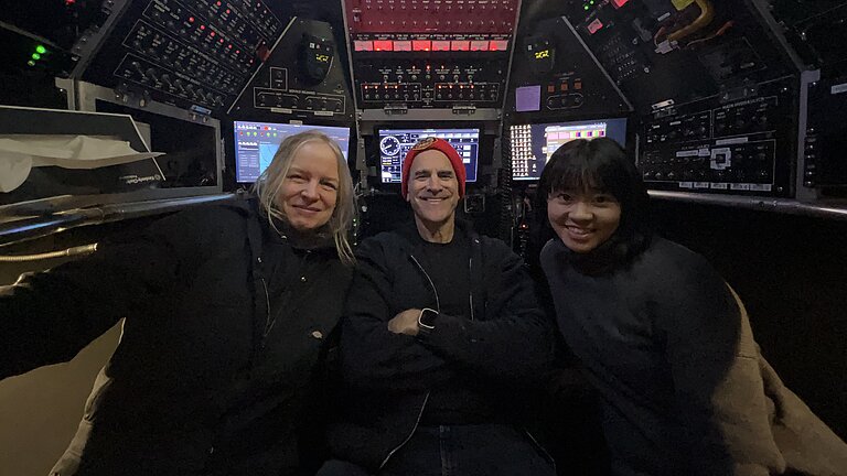 Drei Personen sitzen im Tauchboot ALVIN. Um sie herum Schaltknöpfe und Displays. Sie lachen in die Kamera.
