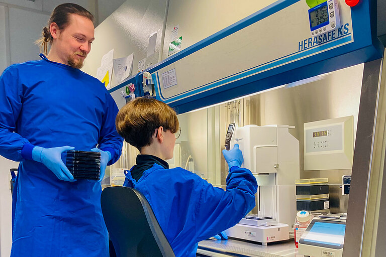 Zwei Mitarbeitende in einem Labor, eine sitzt, einer steht, beide tragen leuchtend blaue Kittel