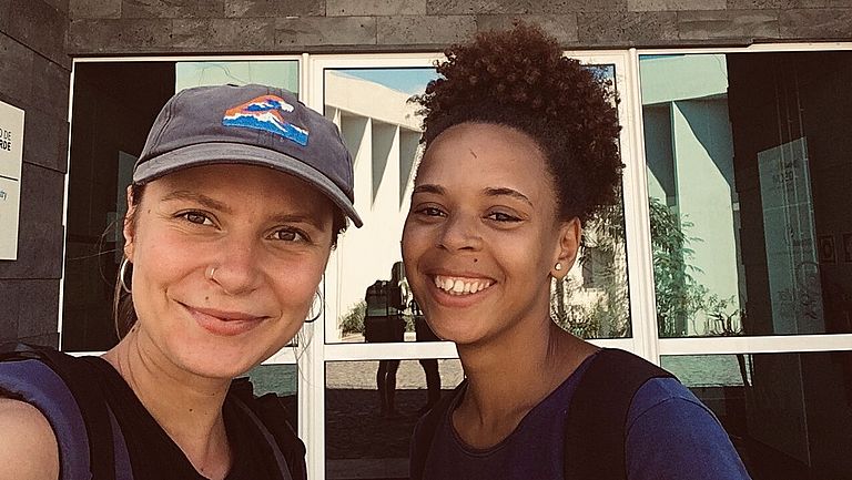 Zwei junge Frauen machen ein Selfie vor einem Gebäude mit der Aufschrift "Ocean Science Center Mindelo"