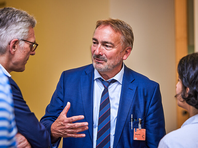 Ein Mann mit kurzen angegrauten Haaren und Dreitagebart im Gespräch mit einem Mann und einer Frau