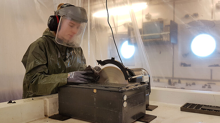 On board the METEOR, a scientist cuts through a rock sample with a circular saw. She wears ear protection and a face shield.