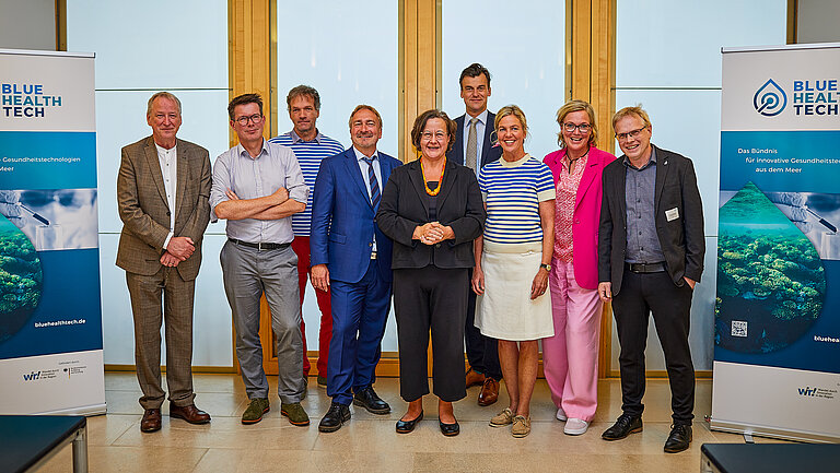 Eine Gruppe von Männern und Frauen in sommerlicher Kleidung, rechts und links stehen Banner, auf denen BlueHealth zu lesen ist.