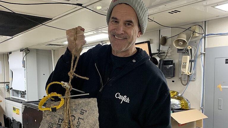 Alvin Pilot Bruce Strickrott bereitet die Ausbringung des Gedenksteins mit dem Tauchboot Alvin vor. Er steht an einem Tisch und hält den Stein in die Kamera. 