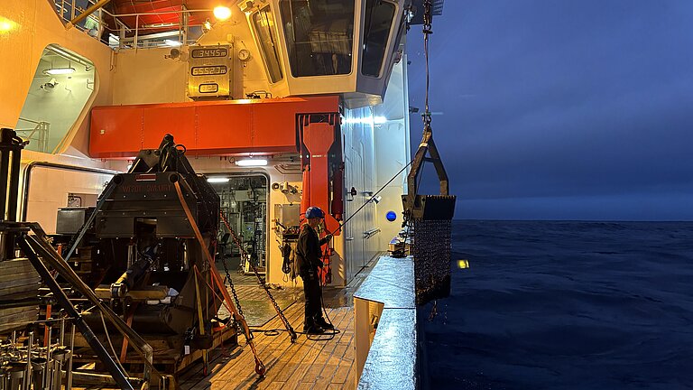  A night-time scene on a research vessel. A person is standing on board the vessel, taking delivery of the dredge that has been hauled out of the water.