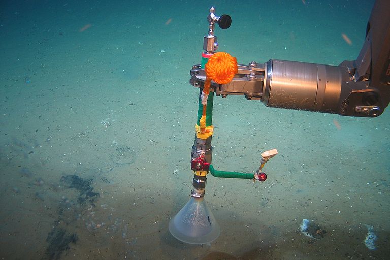 [Translate to English:] Ein Roboterarme hält ein Gerät zur Gasblasenvermessung über einen schlammigen Meeresboden. 