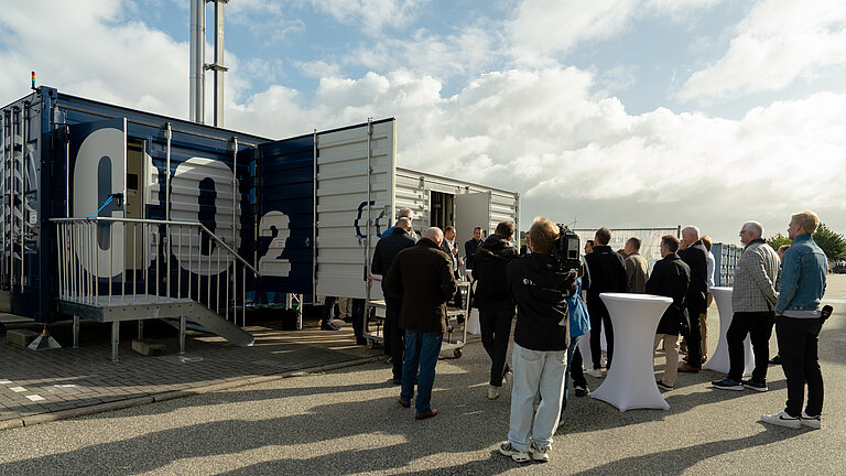 Ein Container mit der Aufschrift "CO2", davor eine Gruppe Männer, teilweise mit Kameras