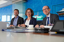 Three persons sit at a table with pens in their hands
