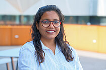A young woman with a dark complexion and long black hair; she is wearing glasses and smiling warmly at the camera