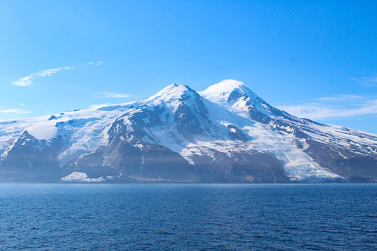 Bild von eisbedeckten Bergen auf einer Insel, davor das Meer, der Himmel darüber ist blau.