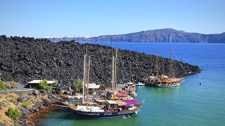 Sailing boats are moored in a small natural harbour of black lava rock. The sea is turquoise blue.