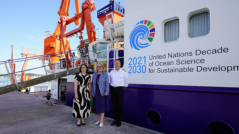 Three people – two women in summer clothes and a man in uniform – are standing in front of a research vessel in the harbour. The gangway has been lowered.