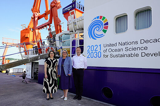 Three people – two women in summer clothes and a man in uniform – are standing in front of a research vessel in the harbour. The gangway has been lowered.