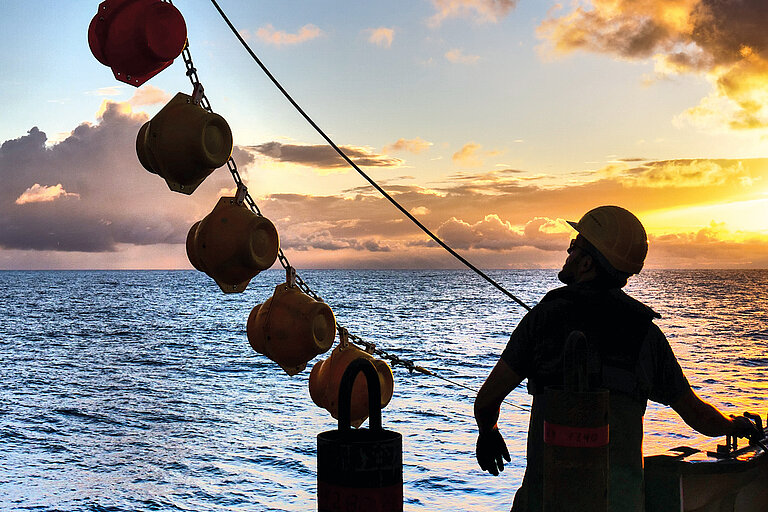 Mann mit Helm an Bord eines Schiffes im Gegenlicht bei Sonnenuntergang. Eine Kette mit großen runden Auftriebskörpern hängt schräg über ihm.