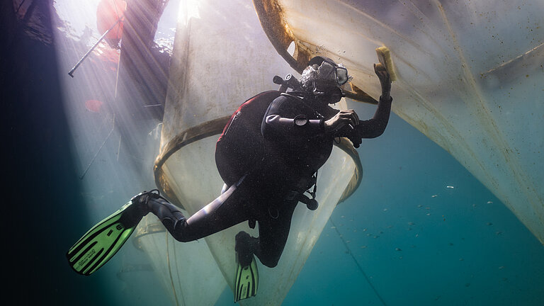 Unterwasserbild: Ein Taucher mit einer Bürste in der Hand schwimmt an großen transparenten Kunststoffsäcken entlang, um sie zu reinigen. 