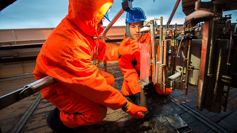 Zwei Männer in leuchtend orangen Overalls an Bord eines Schiffes, zwischen sich eine transparente Röhre, je zur Hälfte mit Sediment und Wasser gefüllt
