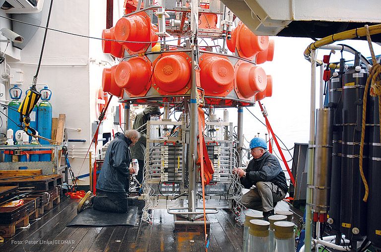 [Translate to English:] Zwei Männer arbeiten an Deck des Schiffs an einem großen Gerät, das im unteren Teil viele Kabel und technische Geräte hat und oben orangene Kugeln, die in Halterungen befestigt sind.