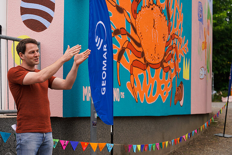 A man in a red T-shirt and jeans is standing in front of a painted wall, gesturing
