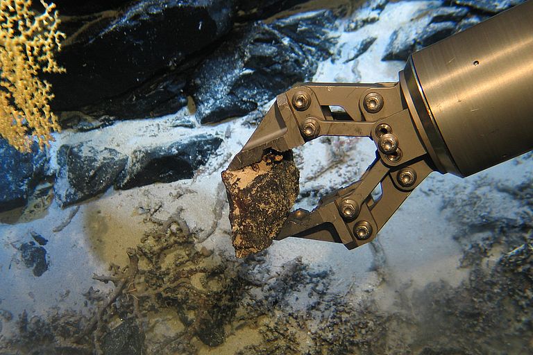 A metal gripper arm grips a piece of rock on the seabed