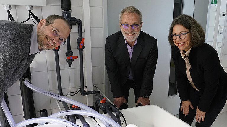 Two men and a woman are standing around white basins with pipes and looking at the camera