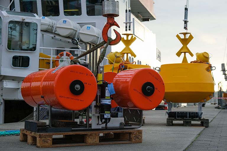 Orange und gelbe Geräte stehen vor dem Forschungsschiff ALKOR auf der Pier. 