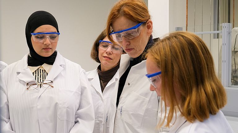 Vier Frauen in weißen Laborkitteln, mit blauen Handschuhen und Schutzbrillen stehen an einem Tisch mit Analysegeräten