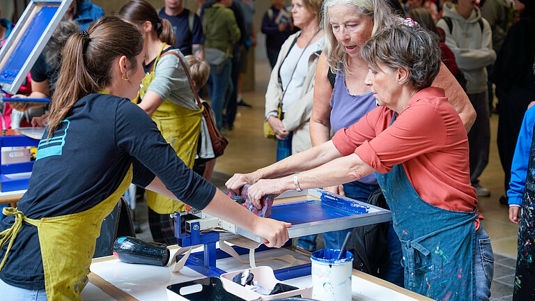 Three women at a screen-printing press; blue ink is being pulled through the screen